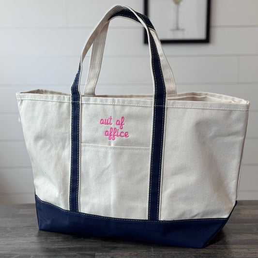 White tote bag with navy accents and 'out of office' text on a wooden surface.