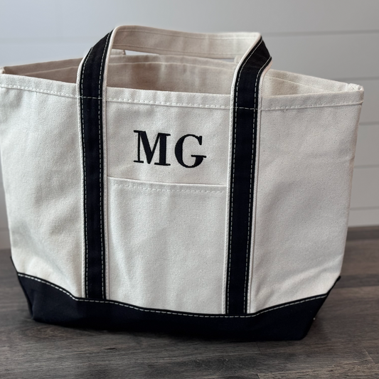 White tote bag with black handles and trim, featuring initials 'MG', on a wooden surface.